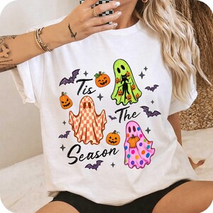 K&ouml;nnte beinhalten: Ein wei&szlig;es T-Shirt mit einer Halloween-Grafik und dem Schriftzug "Tis The Season". Das Design zeigt drei Geister: einen mit Karomuster, einen lindgr&uuml;nen mit einer schwarzen Katze und einen rosa gepunkteten, dazu orangefarbene K&uuml;rbisse und violette Flederm&auml;use.