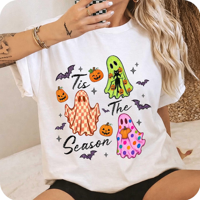 K&ouml;nnte beinhalten: Ein wei&szlig;es T-Shirt mit einer Halloween-Grafik und dem Schriftzug "Tis The Season". Das Design zeigt drei Geister: einen mit Karomuster, einen lindgr&uuml;nen mit einer schwarzen Katze und einen rosa gepunkteten, dazu orangefarbene K&uuml;rbisse und violette Flederm&auml;use.