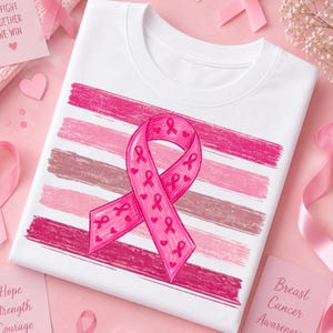 May include: A white t-shirt featuring a large pink awareness ribbon centered over horizontal brushstrokes in varying shades of pink. The ribbon is decorated with smaller pink ribbon icons and heart shapes, creating a layered graphic design.