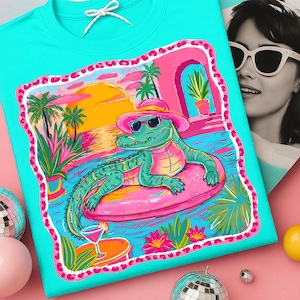 May include: Turquoise t-shirt featuring a colorful graphic of an alligator wearing sunglasses and a pink hat, lounging on a pink float in a pool. The graphic includes palm trees, a sunset, and a cocktail.