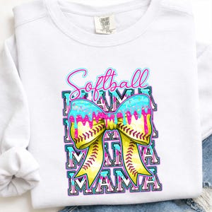 May include: White sweatshirt with a colorful graphic. The design features the words "Softball Mama" in a layered design with a yellow bow, blue icing, and pink drips. The bow has baseball stitching.
