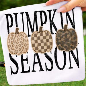 May include: A white t-shirt featuring the black text PUMPKIN SEASON in a distressed font. Three fabric pumpkin appliques are stitched across the centre, displaying a tan leopard print, a brown and tan checkerboard pattern, and a dark camo print.