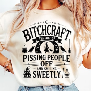 Può includere: T-shirt color crema con un design grafico nero. Il design include il testo "Bitchcraft: The Art of Pissing People Off and Smiling Sweetly" insieme a illustrazioni a tema strega, come una strega, un calderone e corvi.