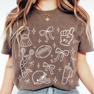 Puede incluir: Una camiseta marr&oacute;n de manga corta con un estampado gr&aacute;fico de l&iacute;neas blancas que muestra una animadora, un bal&oacute;n de f&uacute;tbol americano, un casco, un dedo de espuma con el texto #1, lazos y un vaso, con peque&ntilde;as estrellas dispersas.