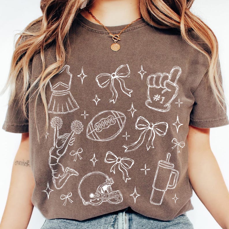 Puede incluir: Una camiseta marr&oacute;n de manga corta con un estampado gr&aacute;fico de l&iacute;neas blancas que muestra una animadora, un bal&oacute;n de f&uacute;tbol americano, un casco, un dedo de espuma con el texto #1, lazos y un vaso, con peque&ntilde;as estrellas dispersas.