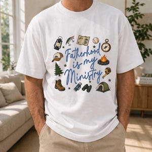 May include: White t-shirt with the text "Fatherhood is my Ministry" in blue script. The shirt features camping-themed illustrations, including a lantern, fishing lure, map, baseball, compass, campfire, hat, pine tree, hiking boot, binoculars, and tent.