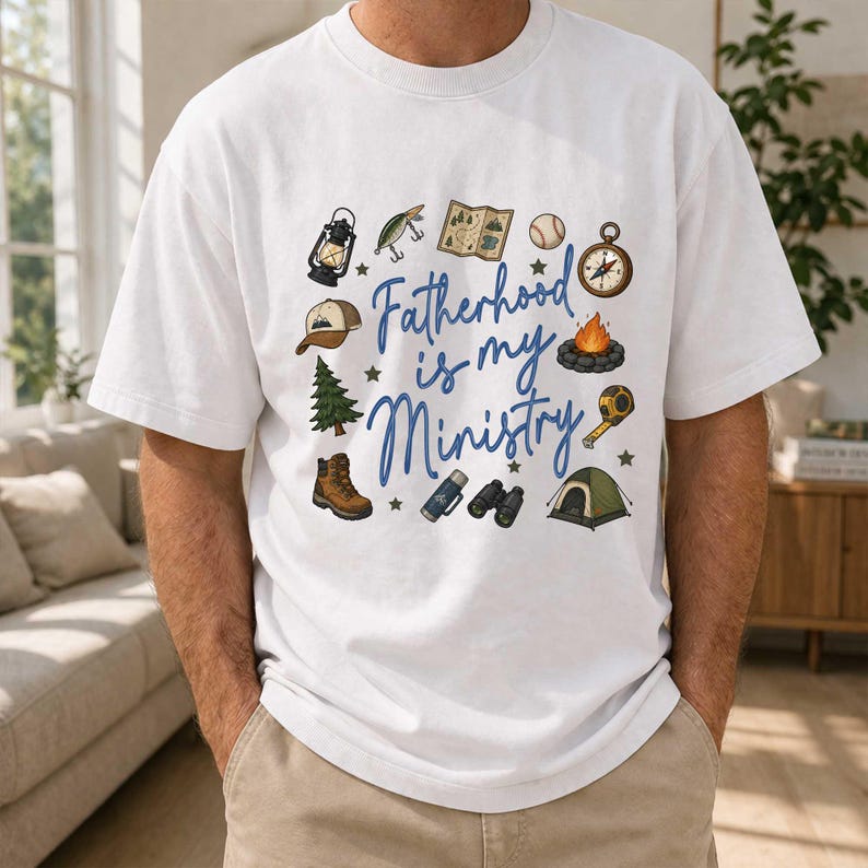 May include: White t-shirt with the text "Fatherhood is my Ministry" in blue script. The shirt features camping-themed illustrations, including a lantern, fishing lure, map, baseball, compass, campfire, hat, pine tree, hiking boot, binoculars, and tent.