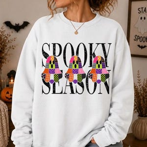 Peut inclure: Un sweat-shirt blanc à manches longues avec le texte noir SPOOKY SEASON et trois motifs en forme de fantômes. Chaque fantôme est composé d'un motif patchwork coloré avec divers designs incluant des pois, des carreaux et des motifs floraux.