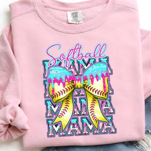 May include: Pink sweatshirt with a graphic that says "Softball Mama." The graphic features a yellow softball bow with blue icing and sprinkles. The word "Mama" is repeated below the bow in a block letter design.