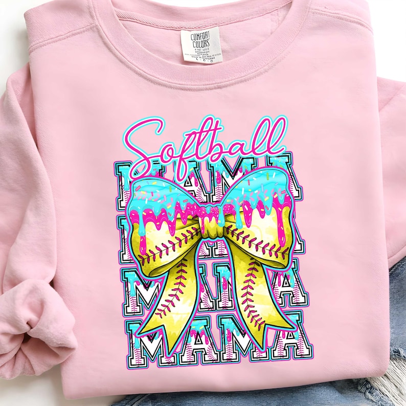 May include: Pink sweatshirt with a graphic that says "Softball Mama." The graphic features a yellow softball bow with blue icing and sprinkles. The word "Mama" is repeated below the bow in a block letter design.