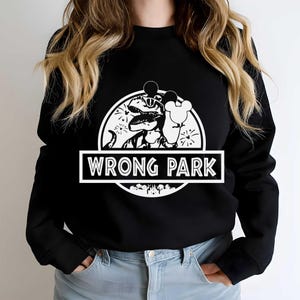 Puede incluir: Sudadera negra con un gráfico blanco que presenta un dinosaurio con gafas de sol y un globo con forma de Mickey Mouse. El texto "WRONG PARK" se muestra en un cuadro rectangular blanco debajo.