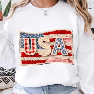 May include: White sweatshirt with a patriotic USA flag design. The flag is made of patchwork fabric with red, white, and blue patterns, including stars and stripes. The word "USA" is prominently displayed across the flag.