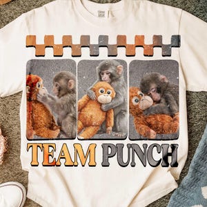 以下が含まれることがあります： クリーム色のTシャツ。3匹の猿のイラストが描かれています。イラストは長方形のフレームに入っており、上部に装飾的なボーダーがあります。「TEAM PUNCH」の文字が下に太字で印刷されています。