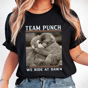 May include: Black t-shirt with the text "TEAM PUNCH WE RIDE AT DAWN" above a black and white image of a monkey hugging a stuffed animal. The shirt has a crew neck and short sleeves.