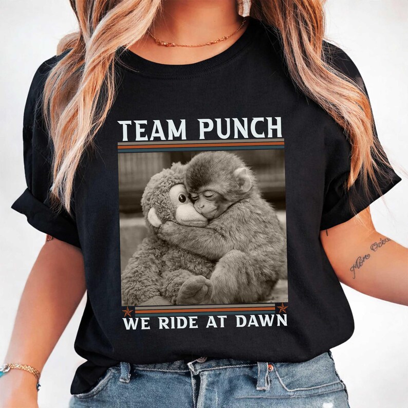 May include: Black t-shirt with the text "TEAM PUNCH WE RIDE AT DAWN" above a black and white image of a monkey hugging a stuffed animal. The shirt has a crew neck and short sleeves.