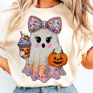 Op de afbeelding: Crèmekleurig T-shirt met een sprankelend spookontwerp met een kleurrijke strik, een pompoenvormige tas en een Halloween-drankje. De geest heeft grote ogen en roze wangen, perfect voor Halloween.