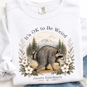 May include: A white long-sleeve sweatshirt featuring a graphic of a raccoon in a forest with a mountain in the background. The text "It's OK to Be Weird" is arched above, and "Jimothy from Seattle" is printed below, framed by white flowers.
