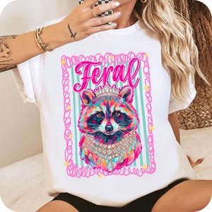 Pu&ograve; includere: T-shirt bianca con un design di procione colorato con corona e gioielli, con la scritta "Feral" in rosa. Il procione &egrave; posto su uno sfondo a righe all'interno di una cornice rosa.