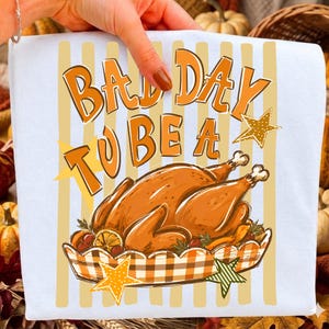May include: A white t-shirt featuring a graphic print with vertical light yellow stripes and the orange text "BAD DAY TO BE A". Below the text is a roasted turkey in a checkered dish, surrounded by orange and green star illustrations.