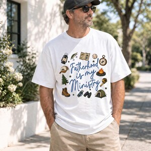 May include: White t-shirt with the text "Fatherhood is my Ministry" in blue script, surrounded by camping-themed illustrations. Includes a compass, tent, campfire, and hiking boots.