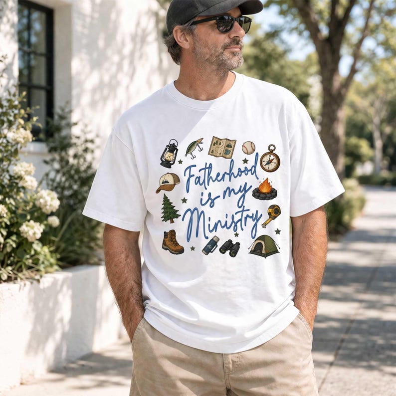 May include: White t-shirt with the text "Fatherhood is my Ministry" in blue script, surrounded by camping-themed illustrations. Includes a compass, tent, campfire, and hiking boots.