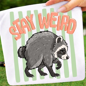 May include: A white fabric pouch featuring a cartoon raccoon illustration in shades of gray and black. Above the raccoon, the text "STAY WEIRD" is printed in a bold, peach-colored font. The background includes vertical light green stripes.