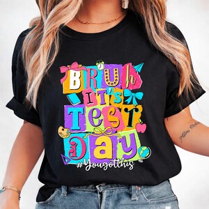 May include: Black t-shirt with a colorful graphic that reads "BRUH IT'S TEST DAY" in a layered, block-letter style. The design includes bows, an alarm clock, an apple, and a globe. The hashtag "#YouGotthis" is at the bottom.