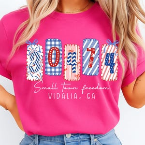 May include: A hot pink t-shirt featuring a graphic design with the numbers "30474" in a patriotic style. The design includes red, white, and blue patterns and the text "Small Town freedom VIDALIA, GA".