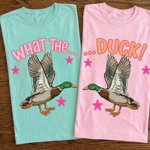 Puede incluir: Dos camisetas, una aguamarina y otra rosa, cada una con un gráfico de pato. La camiseta aguamarina dice "WHAT THE..." en rosa, y la camiseta rosa dice "...DUCK!" en naranja. Ambas camisetas presentan un pato volador y estrellas rosas.