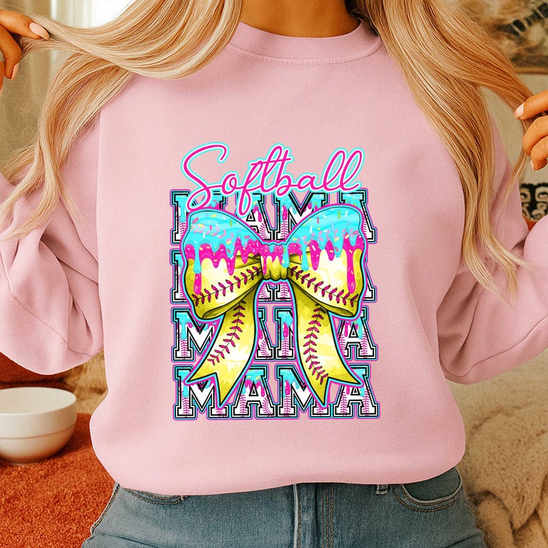 May include: A light pink sweatshirt with a colorful graphic. The graphic features the words "Softball Mama" in a bow design with a softball and dripping icing effect. The sweatshirt is a casual style.