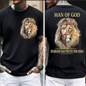 May include: Black t-shirt featuring a split image of Jesus and a lion, with a crown of thorns. The back of the shirt displays the text "MAN OF GOD" above the image and "HUSBAND-DAD-PROTECTOR-HERO" below.