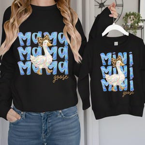 May include: Black sweatshirts with matching designs. One sweatshirt says "Mama Goose" in blue bubble letters with a watercolor goose wearing a bow. The other says "Mini Goose" with the same design. Both have the word "goose" in script.