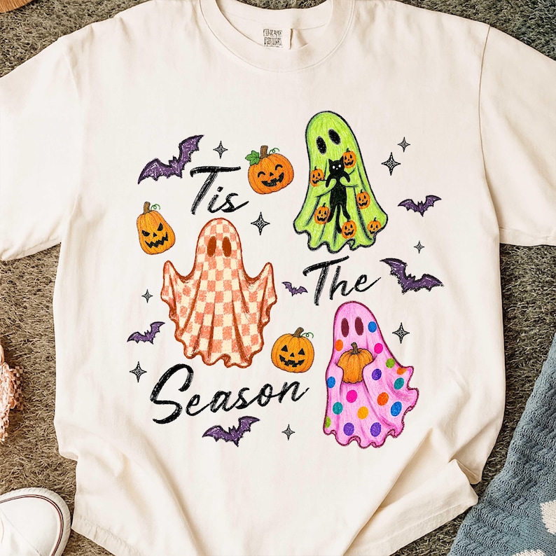 K&ouml;nnte beinhalten: Ein cremefarbenes T-Shirt mit einem Grafikdruck und dem Text 'Tis The Season'. Das Design zeigt drei Geister: einen lindgr&uuml;nen mit einer schwarzen Katze, einen orange-wei&szlig; karierten und einen rosa Geist mit bunten Punkten, dazu K&uuml;rbisse und Flederm&auml;use.