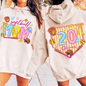 Könnte beinhalten: Cremefarbener Hoodie mit Softball-Motiv. Auf der Vorderseite stehen die Worte "softball MOM" in bunten Buchstaben mit Baseball-Grafiken. Auf der Rückseite befindet sich ein Baseball-Diamant-Design mit der Nummer "20" und dem Namen "Oliver".