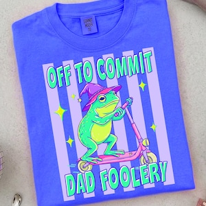 May include: A blue t-shirt with a graphic of a green frog wearing a purple wizard hat and riding a pink scooter. The text "OFF TO COMMIT DAD FOOLERY" is printed on the shirt in a playful font. The shirt has a tag that says "GORNOT HOODER 10".