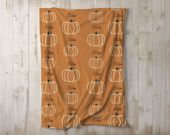 May include: Orange blanket featuring a repeating pattern of white pumpkin outlines and the name "Ellie" in black script. The blanket is displayed against a light wood background, suggesting a fall or harvest theme.