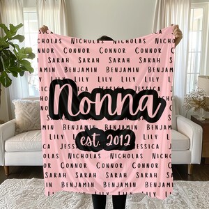 May include: A pink blanket with the word "Nonna" in large black script. The blanket is covered with names in black and the text "est. 2012". A cozy and personalized gift.