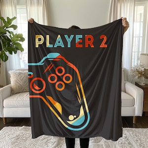 May include: Black blanket with a colorful graphic of a video game controller and the words "PLAYER 2" in orange, blue, and yellow. The blanket is held up in front of a window and a white couch.