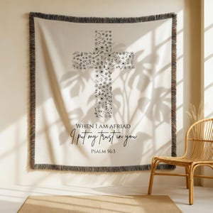 Puede incluir: Manta tejida de color crema con un diseño de cruz hecho de ilustraciones florales. La manta tiene un borde de flecos negros y presenta el texto "WHEN I AM AFRAID I put my trust in you" y "PSALM 56:3".