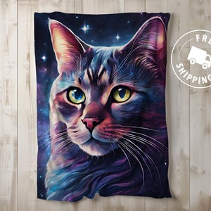 May include: A plush throw blanket featuring a vibrant, artistic rendering of a cat's face against a starry galaxy backdrop. The cat's fur is a mix of purple, blue, and pink hues, with striking yellow and blue eyes. The blanket is displayed on a light wood surface.