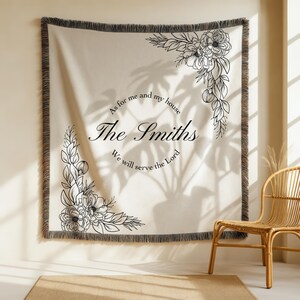 May include: Cream-coloured woven throw blanket with black floral corner designs and fringe trim. The blanket features the text "As for me and my house The Smiths We will serve the Lord" in black script. The blanket is hanging on a wall.