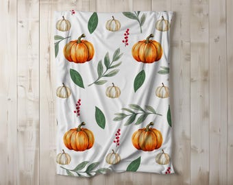 May include: White blanket with a repeating pattern of watercolor pumpkins, green leaves, and red berries. The pumpkins are orange and cream-colored, creating a fall or autumn theme. The blanket is draped over a light wood surface.