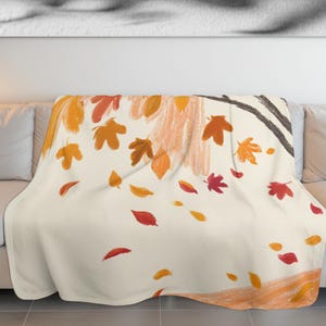May include: A cream-colored throw blanket with a fall-themed design. The blanket features falling leaves in shades of orange, red, and yellow, with a tree branch and trunk in the background. The design is a watercolor style.