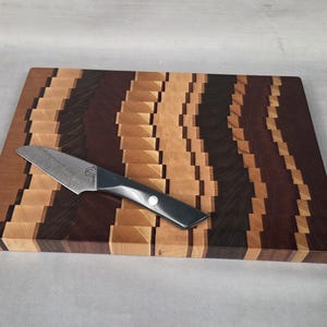 May include: A rectangular wooden cutting board with a zig-zag pattern in shades of brown and tan. A small, black-handled knife with a silver blade rests on the board. The cutting board is approximately 16 inches long and 12 inches wide.