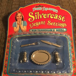 May include: A plastic package with a red background and a picture of a young girl. The package contains a silver toy place setting with a plate, fork, knife, and two salt and pepper shakers. The package says "Silvercast Elegant Settings Non-Tarnishable Metal" and "Imperial Toy Corporation".