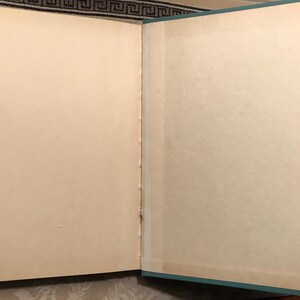 Puede incluir: Un libro abierto con p&aacute;ginas de color crema envejecidas. La encuadernaci&oacute;n es visible, y los bordes de las p&aacute;ginas muestran una ligera decoloraci&oacute;n. La cubierta es de color verde azulado apagado, enmarcando las p&aacute;ginas. El libro parece ser vintage o antiguo.
