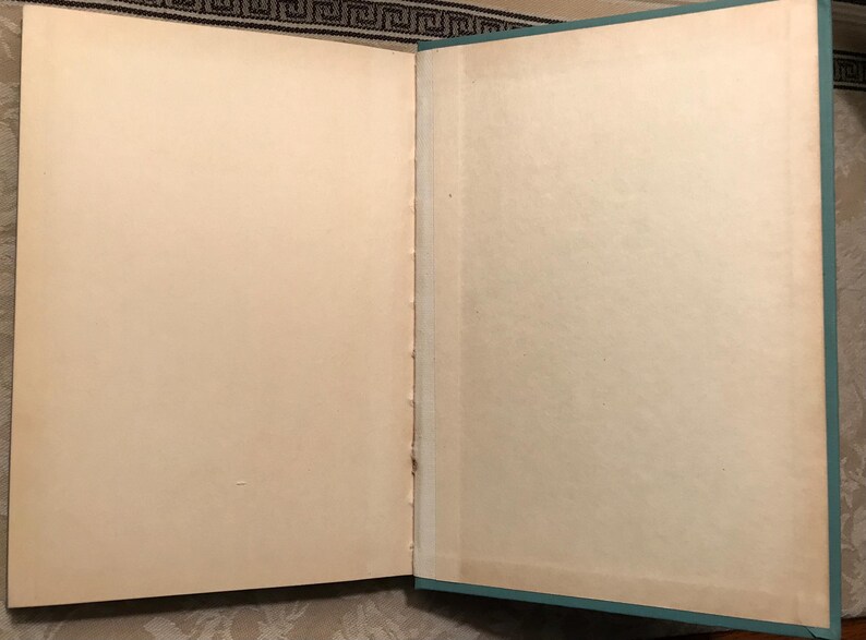 Puede incluir: Un libro abierto con p&aacute;ginas de color crema envejecidas. La encuadernaci&oacute;n es visible, y los bordes de las p&aacute;ginas muestran una ligera decoloraci&oacute;n. La cubierta es de color verde azulado apagado, enmarcando las p&aacute;ginas. El libro parece ser vintage o antiguo.