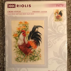 May include: A RIOLIS 1479 cross-stitch kit featuring a rooster standing among yellow and white flowers. The packaging displays the text RIOLIS, 1479, CROSS-STITCH, STRANDED COTTON, and dimensions of 30 x 40 cm or 11 3/4 x 15 3/4 inches.