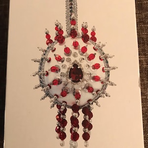 May include: A white box featuring a detailed illustration of a beaded ornament. The ornament has a central teardrop-shaped red gem, surrounded by red beads and silver accents. The box has the text "DWC # 6444" and "MM 202501OR".