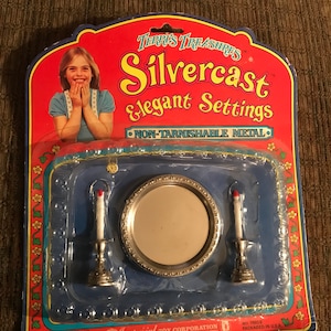 May include: A plastic package with a red, white, and blue border. The package contains a silver-colored toy mirror and two candlesticks. The package has the text "Terris Treasures Silvercast Elegant Settings Non-Tarnishable Metal" on the front. The package also has the text "Imperial Toy Corporation Los Angeles California 90001" on the bottom.